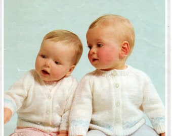 May include: Two babies wearing white knitted cardigans with blue trim. The cardigan has a button closure. The babies are sitting on a light blue surface.