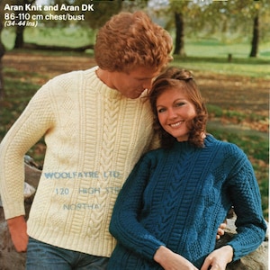 May include: Two people wearing cable knit jumpers. The man is wearing a cream jumper and the woman is wearing a teal jumper. The jumpers have a similar cable knit pattern. The man is wearing blue jeans and the woman is wearing beige trousers. The background is a blurry image of a park.