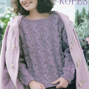 May include: A woman wearing a purple cable knit sweater with a pink coat over it. The text "LEARNING THE ROPES" is at the top of the image. The text below the image reads "Use a pretty tweed yarn for this sweater that is embossed with bold cables. Worked in crochet it will be ready to wear in no time at all."