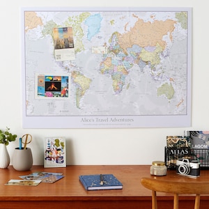 May include: A large world map with travel mementos pinned to it, mounted on a white wall. A desk below holds a notebook, postcards, a camera, and a wire basket with books. The map is labelled "Alice's Travel Adventures."