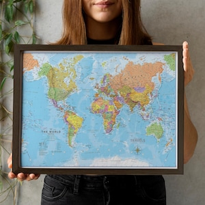 May include: A framed world map in a dark brown wooden frame. The map displays countries in various colours. The text "THE WORLD POLITICAL" and "TRAVELS" are visible. The map is held by a person.