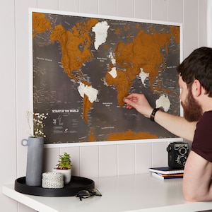 Könnte beinhalten: Eine Person kratzt eine Weltkarte aus Goldfolie ab, um die Länder zu enthüllen, die sie besucht hat. Die Karte befindet sich an einer grauen Wand und trägt den Text "Scratch the World" in weißen Buchstaben.