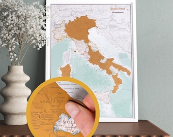 Könnte beinhalten: Eine gerahmte Rubbelkarte von Italien mit weißem Rahmen. Die Karte hat einen hellen Hintergrund, wobei die Regionen mit goldfarbener Folie bedeckt sind. Ein kreisförmiger Ausschnitt zeigt eine Hand, die mit einer Münze die Folie abkratzt.