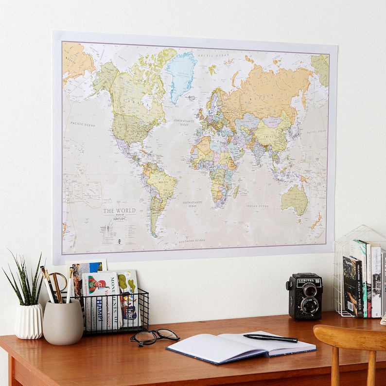 May include: A world map poster with a beige background and colorful country borders hangs on a white wall above a wooden desk. The desk has a white ceramic vase with a green plant, a black wire basket with books, a pair of glasses, a blue notebook, a black pen, and a vintage camera.