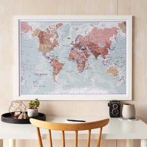 May include: A framed world map with a white frame and a light blue background. The map is in a vintage style with a pink and brown colour scheme. The map is hanging on a light wood wall. There is a desk in the foreground with a camera, a vase, and a plant.