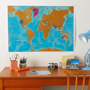 Könnte beinhalten: Eine rechteckige Wandkarte mit dem Titel SCRATCH THE WORLD, die einen leuchtend blauen Ozeanhintergrund und goldfarbene Landmassen zeigt. Einige Regionen sind freigerubbelt, um farbenfrohe Details zu enthüllen. Die Karte hängt an einer weißen Wand.