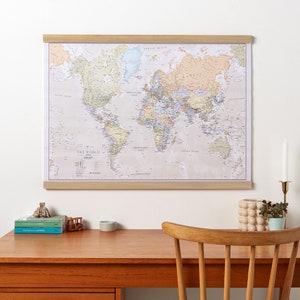 May include: A framed world map with a beige background and colorful country borders. The map is hanging on a white wall above a wooden desk with a chair, a small potted plant, and a few books.