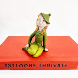 May include: A small figurine of a seated elf with light brown hair, wearing a green pointed hat, a green long-sleeved tunic with a row of buttons, and light green pants. The elf has pointed ears and is positioned on a bright red book.