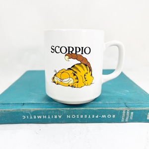 May include: White ceramic mug featuring a cartoon Garfield character with the word "SCORPIO" above it. The mug has a handle and is resting on a vintage blue book. The Garfield character is yellow and orange.