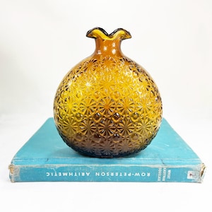 May include: Amber-colored glass vase with a textured, starburst pattern. The vase has a ruffled rim and a rounded body. It sits on a vintage blue book. The vase is a decorative item.