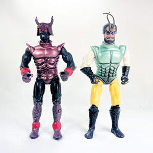 May include: Two action figures stand side-by-side. One is a dark figure with a horned helmet, red accents, and black limbs. The other figure has a green chest, yellow legs, and black boots. Both are vintage toys.