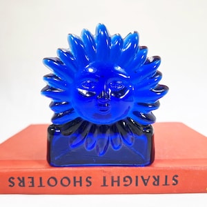 May include: A vibrant cobalt blue glass sun figurine featuring a molded face with radiating sunbeams. The sun sculpture is positioned on top of a rectangular orange book with the text "STRAIGHT SHOOTERS" and "Webster" and "Osborn Riefling" printed on the spine.