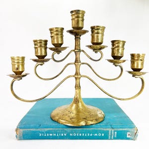 May include: A seven-arm brass candelabra with a central stem and curved arms, each topped with a cylindrical candle cup. The candelabra rests on a teal-colored book titled Row-Peterson Arithmetic, which features white text on its spine.