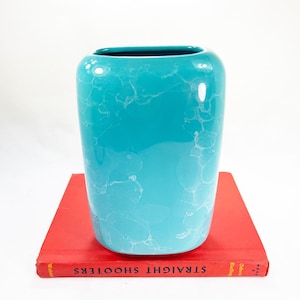 May include: A glossy turquoise ceramic vase with a rounded rectangular shape and a white marbled or crystalline glaze pattern. The vase is positioned on top of a red book titled Straight Shooters, which has visible black text on the spine.