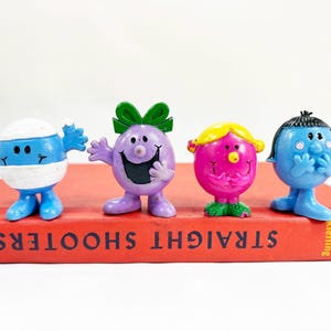 May include: Four small, round, colorful plastic character figurines standing on a red book titled STRAIGHT SHOOTERS. The figures include a blue one with a white bandage, a purple one with a green bow, a pink one with yellow hair, and a blue one.