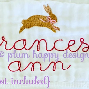 May include: A white embroidered baby blanket with the name "Frances Ann" in red cursive script and a brown bunny with a pink bow above the name. The text "&copy; plum happy designs" is below the name.