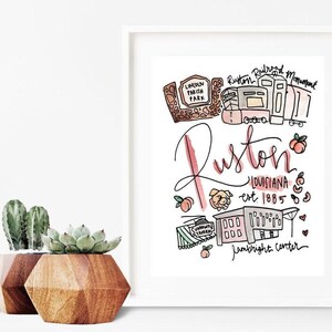 May include: A framed watercolor print featuring the word "Ruston" in cursive, with illustrations of local landmarks and the text "Louisiana est. 1885." The print includes images of peaches, a dog, and a train.
