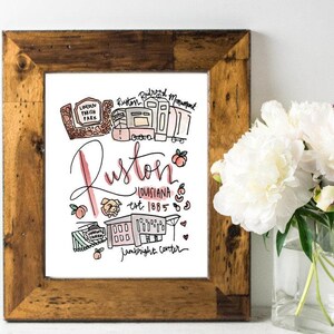 May include: A framed art print with a rustic wooden frame. The print features a hand-drawn illustration of Ruston, Louisiana, with landmarks and the text "Ruston, Louisiana est. 1885". Peaches and other small illustrations are included.