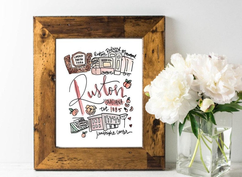 May include: A framed art print with a rustic wooden frame. The print features a hand-drawn illustration of Ruston, Louisiana, with landmarks and the text "Ruston, Louisiana est. 1885". Peaches and other small illustrations are included.