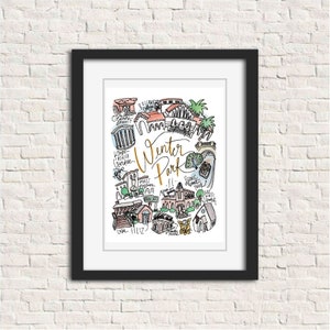 May include: A framed art print featuring a colorful hand-drawn map of Winter Park, Florida. The artwork includes various landmarks and the text "Winter Park, FL" in gold script. The print is in a black frame and is displayed against a white brick wall.