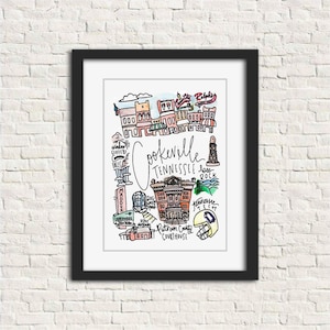 May include: A framed watercolor print of Cookeville, Tennessee, featuring local landmarks and the city's name in cursive. The artwork includes illustrations of buildings, a lighthouse, and a football helmet, all in a colorful, hand-drawn style.
