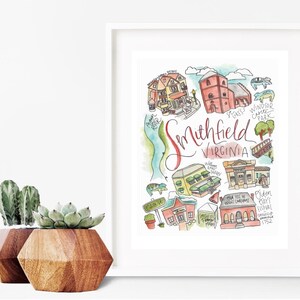 May include: A framed watercolor print of Smithfield, Virginia, featuring local landmarks and the town's name in cursive. The artwork includes illustrations of buildings, a river, and park, all in vibrant colors. Two geometric wooden planters with succulents are in the foreground.