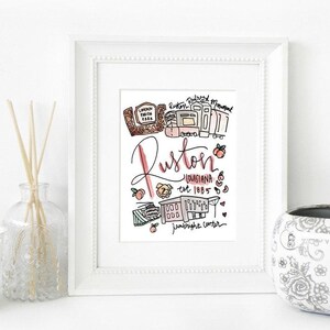 May include: A framed art print featuring a hand-drawn illustration of Ruston, Louisiana. The artwork includes the city's name in cursive, landmarks, and the establishment year, all in a colorful, whimsical style. The print is displayed on a white surface.