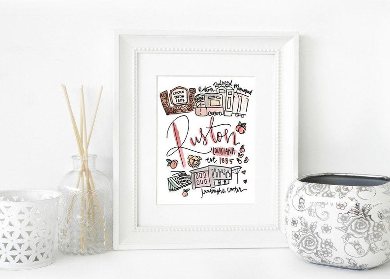 May include: A framed art print featuring a hand-drawn illustration of Ruston, Louisiana. The artwork includes the city's name in cursive, landmarks, and the establishment year, all in a colorful, whimsical style. The print is displayed on a white surface.