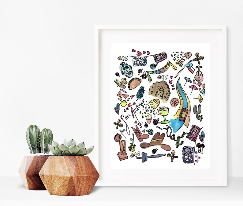 May include: A framed watercolor print featuring colorful illustrations of San Antonio landmarks and cultural symbols. The artwork includes a skull, guitar, river, and the text "San Antonio". Two wooden geometric planters with succulents are in the foreground.