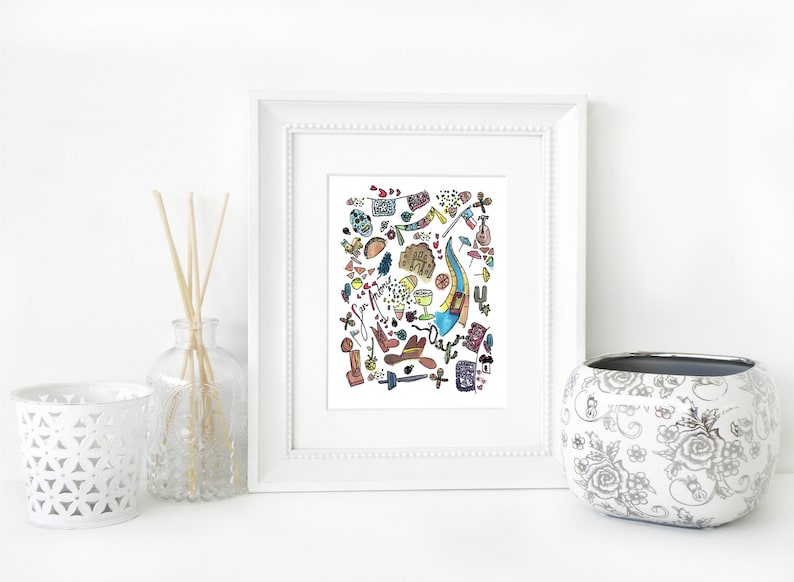 May include: A framed watercolor print featuring a colorful illustration of various objects, including a guitar, cowboy boots, and a skull. The print is displayed on a white surface with decorative items.