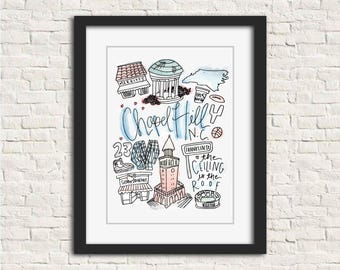 May include: A framed art print with a hand-drawn illustration of Chapel Hill, North Carolina. The artwork includes local landmarks, the text "Chapel Hill N.C.", and other icons. The print is displayed on a white brick wall.
