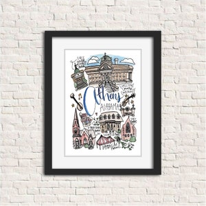 May include: A framed art print featuring a colorful, hand-drawn illustration of Athens, Alabama. The artwork includes various landmarks and the text "Athens Alabama" in a stylized font. The print is set against a white background.