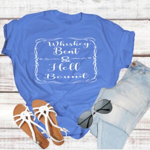 May include: A blue t-shirt with the text "Whiskey Bent & Hell Bound" in a white decorative frame. The image also includes a pair of light blue jeans, sunglasses, and sandals with white straps and brown soles.