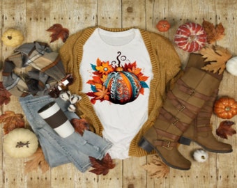May include: A fall-themed flat lay featuring a white t-shirt with a pumpkin graphic, a mustard-colored cardigan, blue jeans, brown boots, a plaid scarf, and decorative pumpkins and leaves. The t-shirt has a detailed pumpkin design.