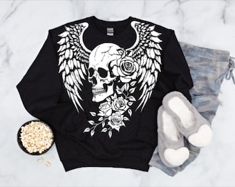May include: Black sweatshirt with a white skull design, wings, and roses. Gray camouflage sweatpants and gray heart-shaped slippers are next to the sweatshirt. A bowl of popcorn is in the lower left corner.