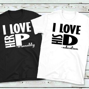 May include: Two matching t-shirts, one black and one white. The black shirt says "I LOVE HER P - personality" in white block letters. The white shirt says "I LOVE HIS D - education" in black block letters. Both shirts are short-sleeved.