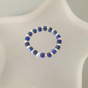 May include: A ring composed of dark blue lapis lazuli beads and silver-coloured beads. The beads are arranged in a continuous band. The ring is displayed on a light, reflective surface.