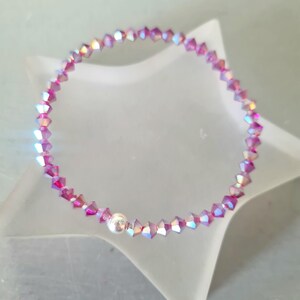 May include: A bracelet made of iridescent pink crystal beads. The bracelet has a silver-coloured clasp and is displayed on a white star-shaped surface. The crystals reflect a range of colours, adding a touch of sparkle.