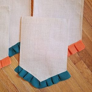 May include: Three burlap banners with different colored ruffled edges. The banner on the left has orange ruffles, the middle banner has teal ruffles, and the banner on the right has orange ruffles.