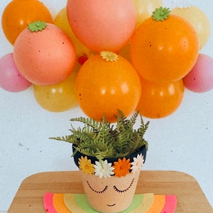 May include: A small terracotta flower pot with a painted face and a flower crown. The pot is sitting on a rainbow-colored paper cutout and surrounded by pastel-colored balloons.