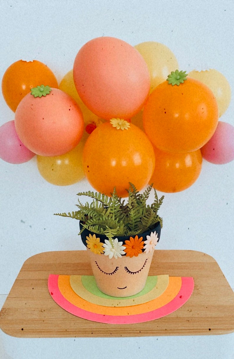May include: A small terracotta flower pot with a painted face and a flower crown. The pot is sitting on a rainbow-colored paper cutout and surrounded by pastel-colored balloons.