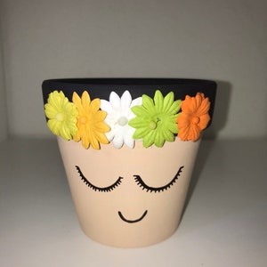 May include: A small terracotta planter with a black rim and a painted face. The planter has a smiling mouth and closed eyes. The rim is decorated with five colorful felt flowers.