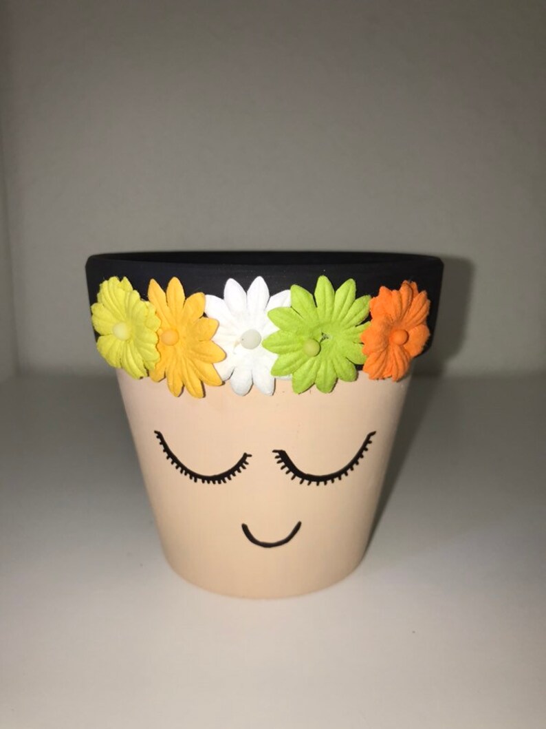 May include: A small terracotta planter with a black rim and a painted face. The planter has a smiling mouth and closed eyes. The rim is decorated with five colorful felt flowers.