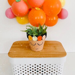 May include: A small terracotta pot with a painted face and a black headband decorated with colorful flowers. The pot is filled with green ferns and is sitting on a white and wood storage bin with a polka dot pattern.