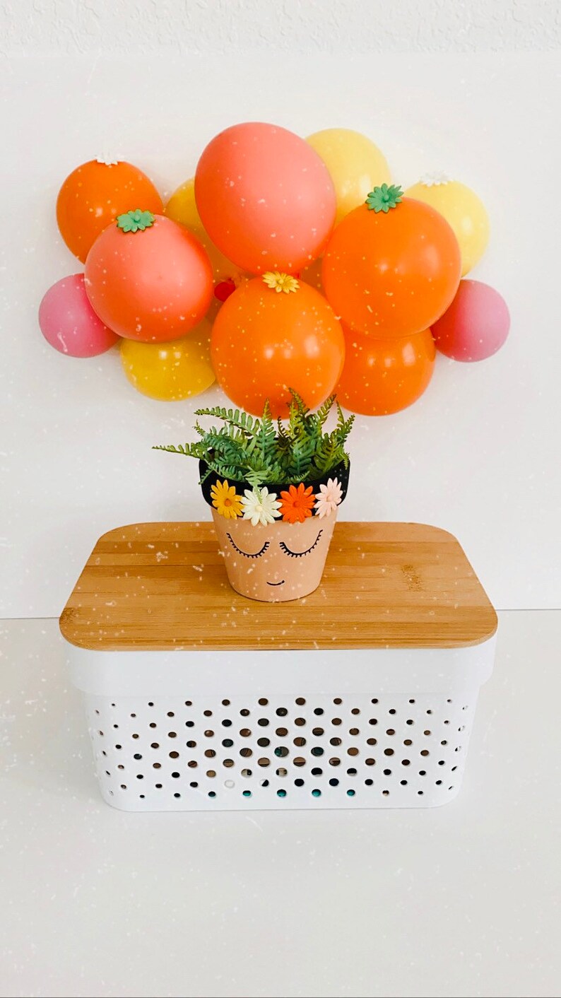 May include: A small terracotta pot with a painted face and a black headband decorated with colorful flowers. The pot is filled with green ferns and is sitting on a white and wood storage bin with a polka dot pattern.