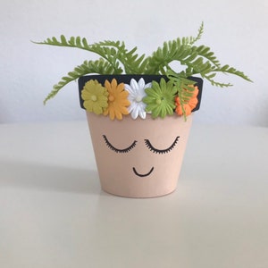 May include: A small terracotta planter with a painted face, closed eyes, and a smile. The planter is decorated with a black rim and a row of colorful felt flowers.