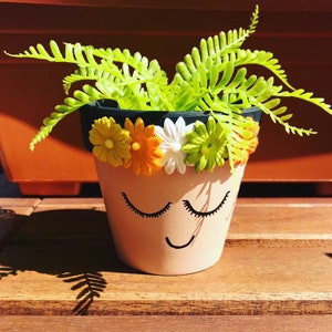 May include: A terracotta flower pot with a painted face and a black rim. The pot is decorated with a colorful flower crown and has a green fern plant growing out of it.