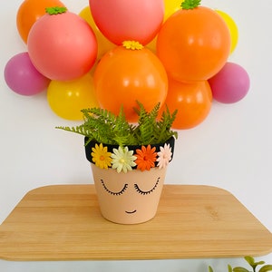 May include: A terracotta flower pot with a painted face and a black headband decorated with colorful flowers. The pot is sitting on a wooden surface with a background of colorful balloons.