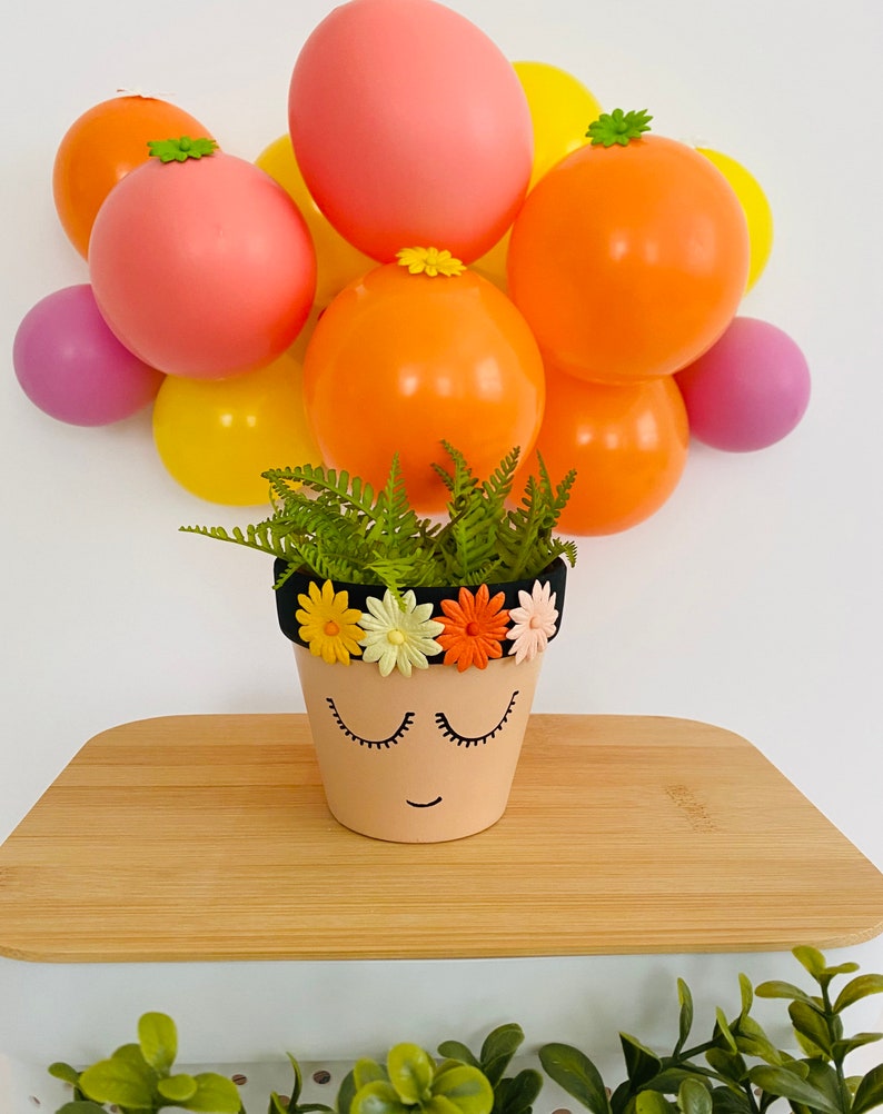May include: A terracotta flower pot with a painted face and a black headband decorated with colorful flowers. The pot is sitting on a wooden surface with a background of colorful balloons.