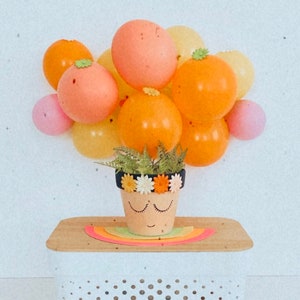 May include: A terracotta flower pot with a painted face and a flower crown is surrounded by pastel balloons in shades of pink, yellow, and orange. The pot is sitting on a wooden surface.